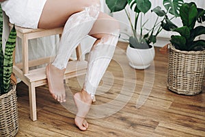 Young woman in white towel applying shaving cream on her legs in home bathroom with green plants. Skin care and wellness. Hair