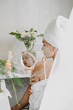 Young woman in white towel chilling in bedroom and making clay facial mask near mirror. Girl doing beauty treatment and