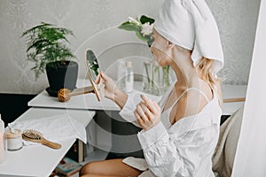 Young woman in white towel chilling in bedroom and making clay facial mask near mirror. Girl doing beauty treatment and