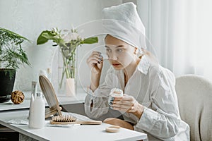 Young woman in white towel chilling in bedroom and making clay facial mask near mirror. Girl doing beauty treatment and