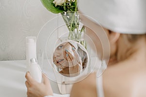 Young woman in white towel chilling in bedroom and making clay facial mask near mirror. Girl doing beauty treatment and