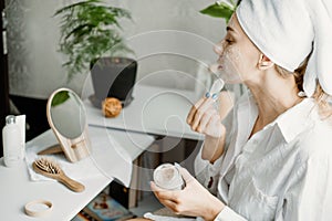 Young woman in white towel chilling in bedroom and making clay facial mask near mirror. Girl doing beauty treatment and