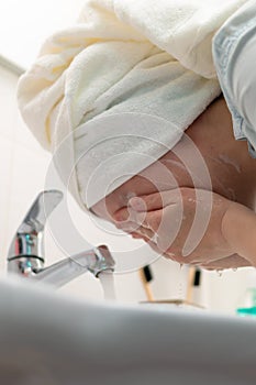 A young woman with a white towel on her head in the bathroom washes off a facial mask. Selective focus
