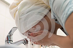 A young woman with a white towel on her head in the bathroom washes off a facial mask. Selective focus