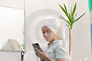 A young woman with a white towel on her head in a bright bathroom communicates with a smartphone