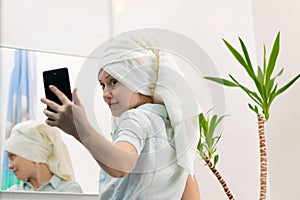 A young woman with a white towel on her head in a bright bathroom communicates with a smartphone