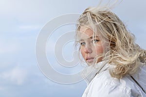 A young woman with wind-blown hair. Pretty blonde girl in a white jacket against a blue clear sky. Close-up