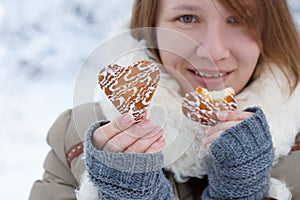 Young woman in winter coat and knitted grey mittens hold beautiful heart shaped biscuit cookies, one bitten, with white icing