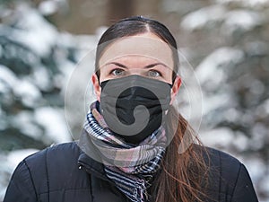 Young woman in winter jacket with black disposable single use face virus mask, blurred snow covered trees background