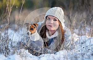 Young woman in winter jacket lying down at snow covered ground, holding her Jack Russell terrier dog, blurred trees background