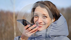 Young woman with wireless headphone is using her smartphone to send a voice message to a new social network Clubhouse standing