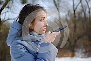 Young woman with wireless headphone is using her smartphone to send a voice message to a new social network Clubhouse standing