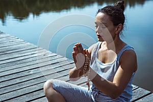 A young woman on a wooden pier sits in a lotus position with her hands folded in a prayer position. Meditation, yoga in nature