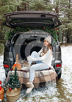 A young woman in a wool hat sits in the trunk of a car and smiles. Winter trip, snow-covered trees in the forest. Snowy