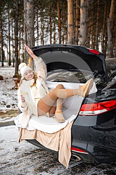 Young woman in woolen hat sits in the trunk of the car. Girl have a fun in black car. Winter travel, snow-covered trees