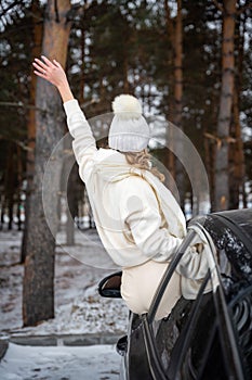 Young woman in woolen white hat have a fun in the car. Winter travel, snow-covered trees in the forest.