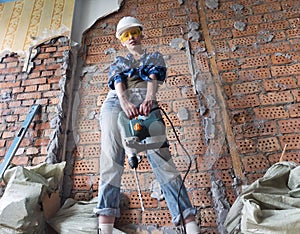 Young woman in work clothes and a protective helmet stands next to a brick wall and holds a heavy hammer drill