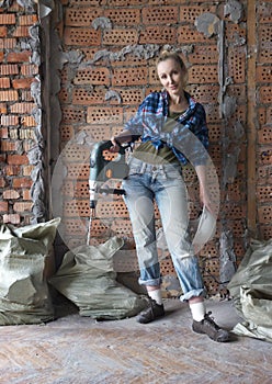 Young woman in work clothes and a protective helmet stands next to a brick wall and holds a heavy hammer drill