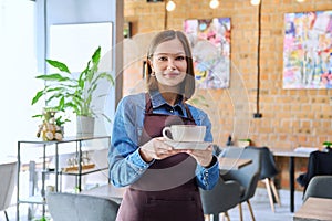 Young woman worker, owner in apron holding cup of coffee in restaurant, coffee shop