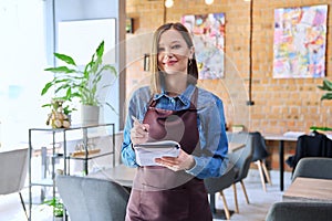 Young woman worker, owner in apron holding notepad pen in restaurant, coffee shop