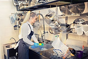 Young woman worker washing dishes using sponge in restaurant sink at end of working day