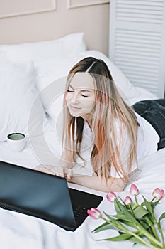 Young woman working on laptop in bed with white linens. Lifestyle composition. Working at home, quarantine concept.