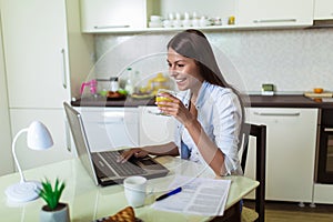 Young woman working with laptop computer and documents while sitting at the kitchen