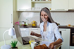 Young woman working with laptop computer and documents while sitting at the kitchen