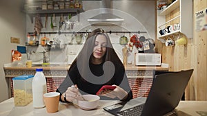 Young woman working laptop computer, using smartphone and eats Corn Flakes Cereal at home kitchen.