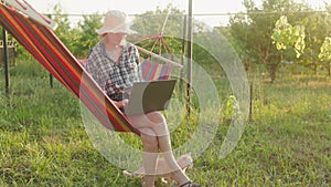 A young woman working on a laptop while lying in a hammock in the garden. Self-isolation, freelancing