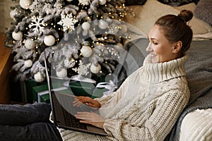 Young woman working or learning on her laptop sitting in living room in quarantine lockdown over Christmas decoration. Social