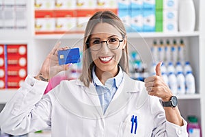 Young woman working at pharmacy drugstore holding credit card smiling happy and positive, thumb up doing excellent and approval