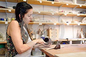 Young woman working raw ceramic and clay to make future ceramic product. Pottery workshop with natural light scraping, smoothing,