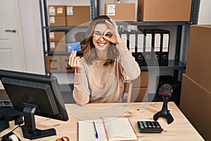 Young woman working at small business ecommerce holding credit card smiling happy doing ok sign with hand on eye looking through