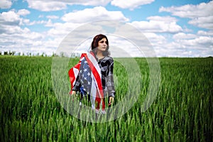 Young woman wrapped in an american flag on the green wheat field. Patriotic holiday celebration. United States of