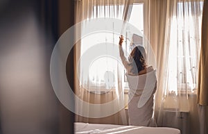Young woman wrapped in a blanket opening the curtains on the window in the morning near the bed