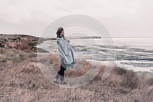 Young woman wrapped in warm blanket is standing on coast of sea in windy weather