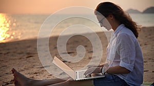 A young woman writer sitting on the beach with her laptop and typing a text.