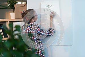 Young woman writing day plan on white board, holding marker in right hand. Student planning schedule rear view portrait