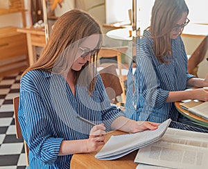 Young woman writing in diary, sitting at wooden desk in coffee shop with notebooks and books