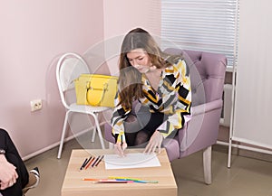 Young woman writing down a note with colour pencils sitting in the armchair at the doctor`s office