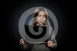 A young woman 25-30 years old holds a text frame on a dark background, shyly averting her eyes to the side photo