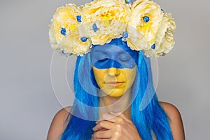 Young woman with yellow blue face art, wreath of peonies on head, holding hands to chest, praying