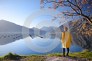 Young woman in yellow coat standing on the coast of Adriatic sea and admiring of stunning winter view of Boka Kotor Bay seascape.