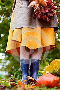 Young woman in yellow dress, cardigan, linen apron, blue rubber boots with bouquet of red maple leaves. Autumn harvest. Cut orange