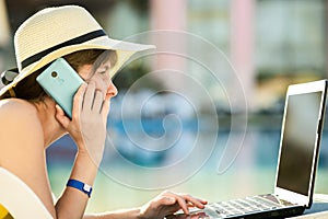 Young woman in yellow dress is laying on beach chair working on computer laptop having conversation on mobile phone in summer