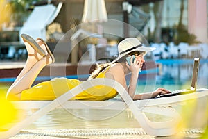 Young woman in yellow dress is laying on beach chair working on computer laptop having conversation on mobile phone in