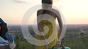 Young woman in yellow dress taking green suitcase from car trunk. Travel and vacations concept.