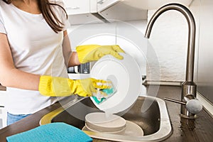 A young woman in yellow gloves washes dishes with a sponge in the sink. House professional cleaning service.