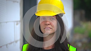 Young woman in yellow hard hat and green high visibility vest, long dark hair, looking to camera, smiling at the end. Blurred